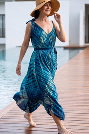 A woman wearing a blue and white tie-dye dress and a wide-brimmed hat walks on a wooden boardwalk near a body of water.