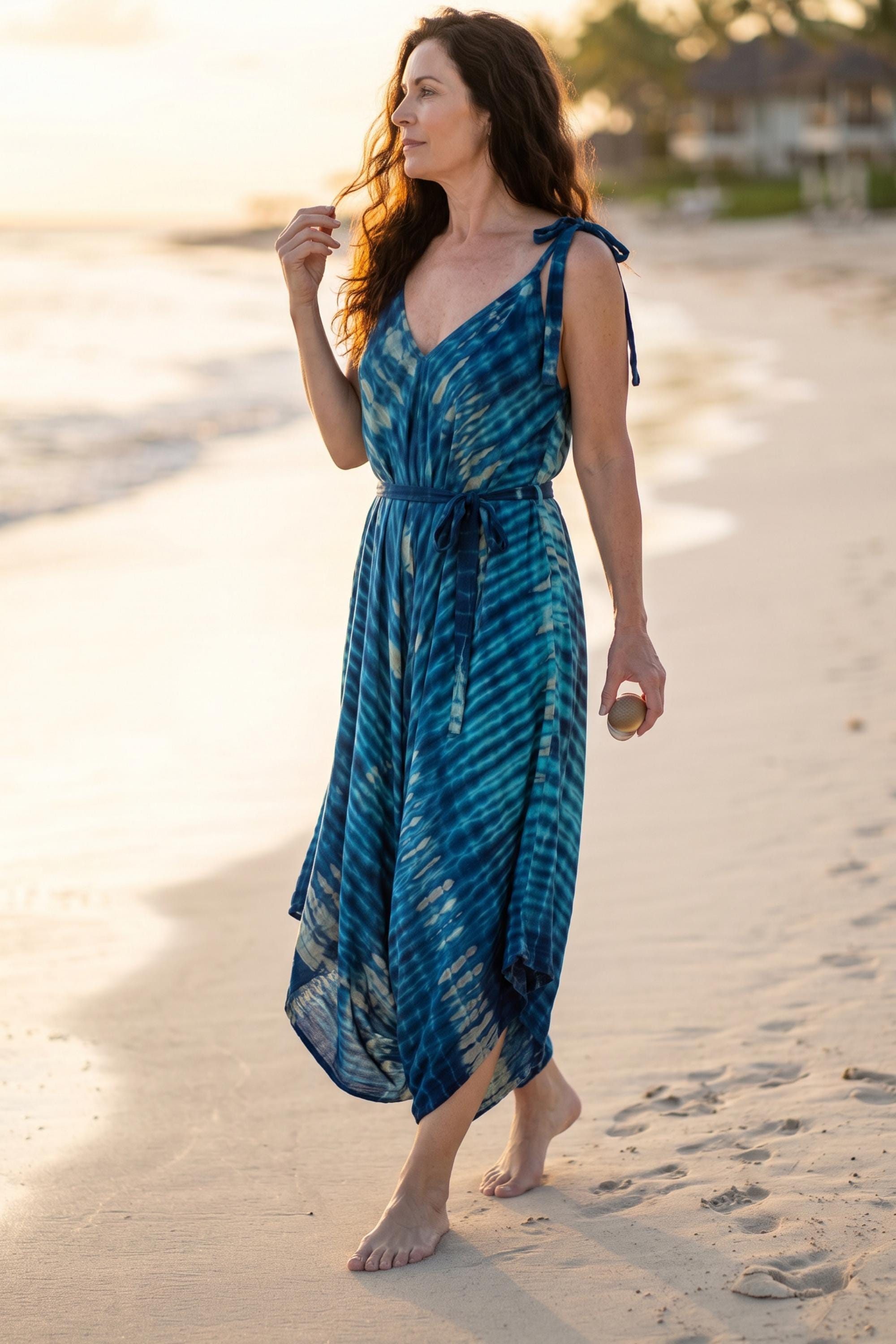 A woman in a blue tie-dye dress walks along a sandy beach, her hair blowing in the wind.