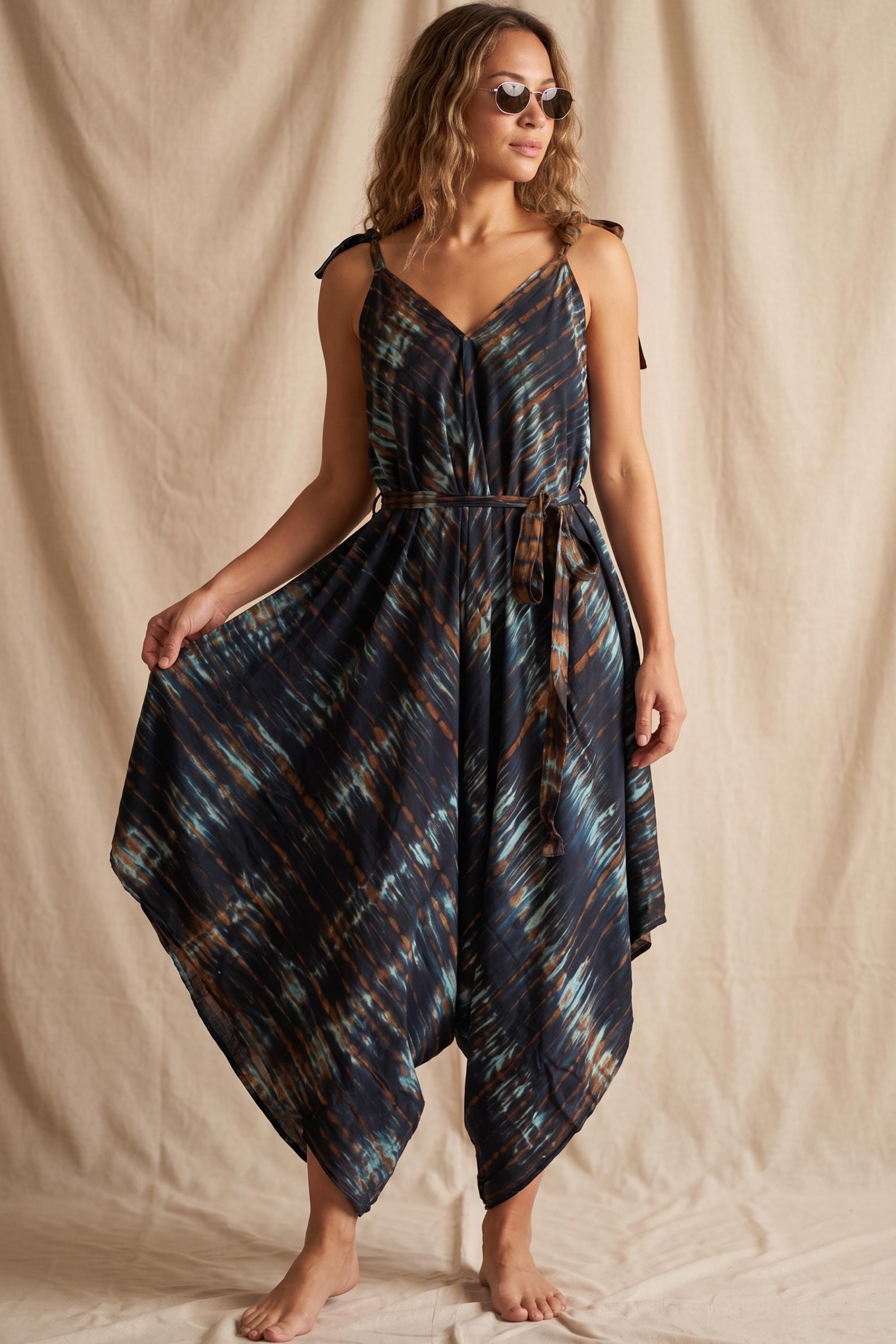 A woman wearing a blue and brown tie-dye dress with a high-low hemline and a tie at the waist, posing against a beige background.