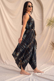 A woman wearing a black and blue tie-dye jumpsuit with a high neckline and a tie at the waist, posing in front of a beige background with palm leaves.