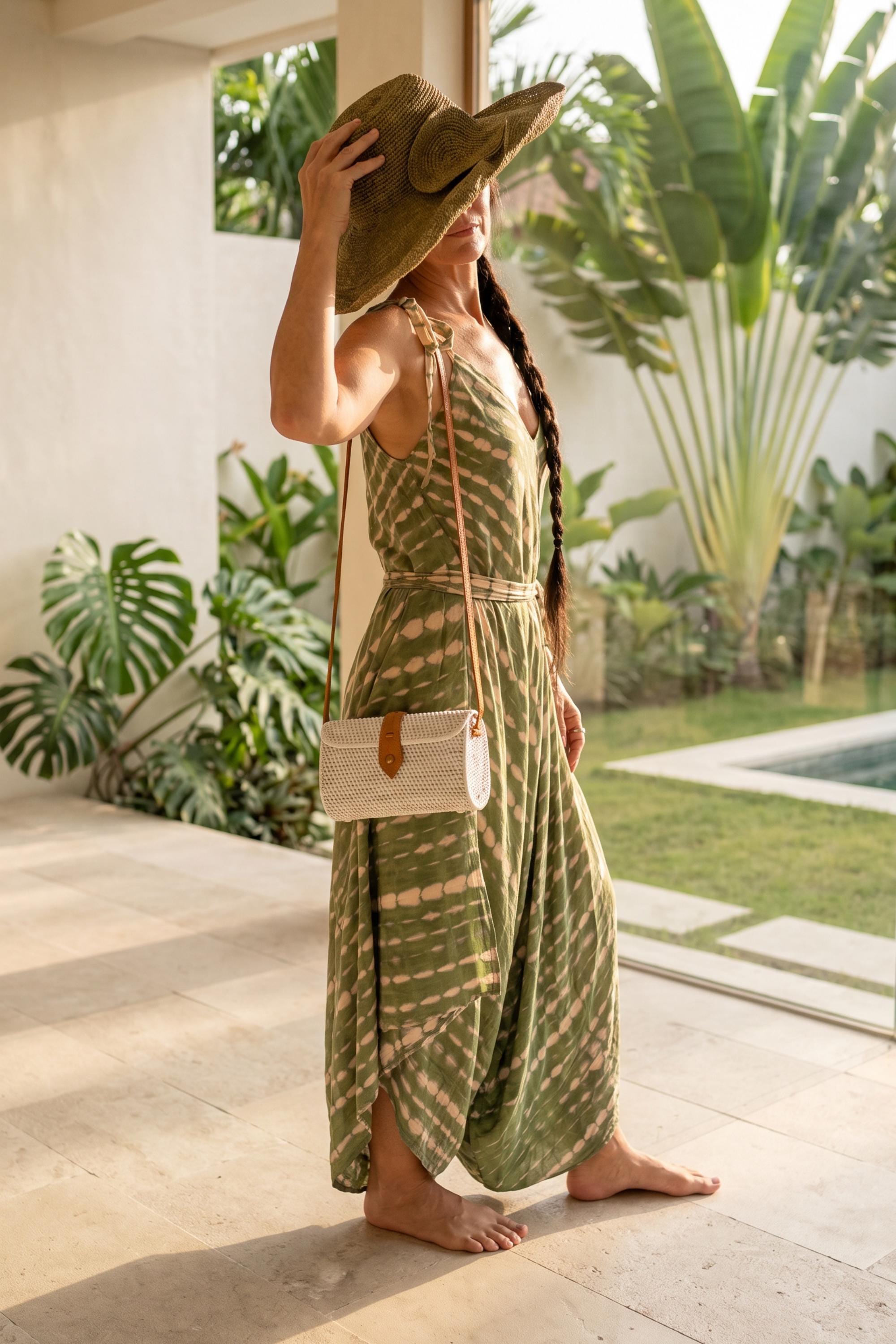A woman in a green and white dress stands in a tropical setting, wearing a wide-brimmed hat and carrying a white purse.