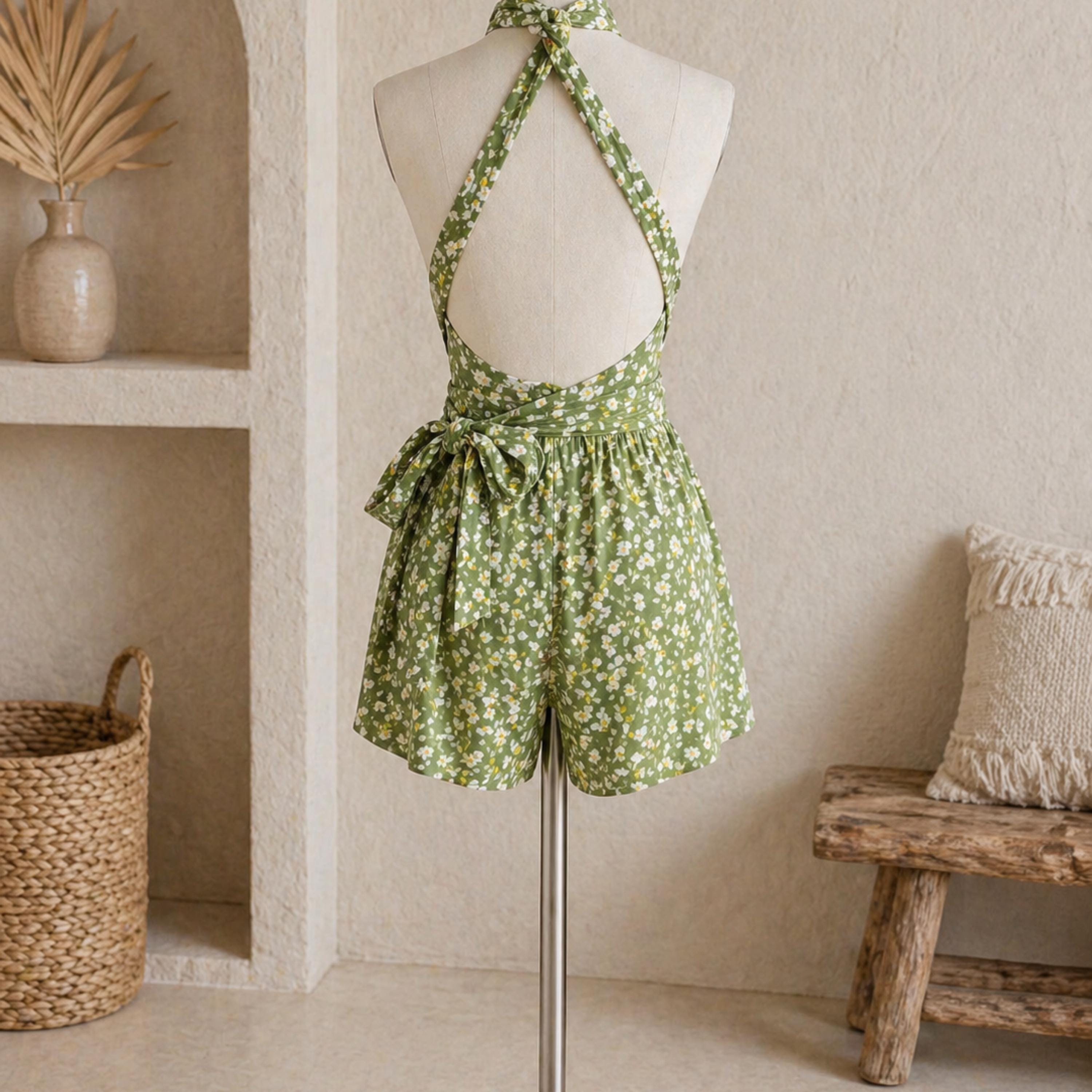 a mannequin wearing a green floral print dress with a high back and a bow at the waist. The dress is displayed on a mannequin stand, and there is a woven basket and a wooden bench in the background.