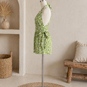 a mannequin wearing a green floral dress with a bow detail, placed in a room with a wooden bench and a woven basket.