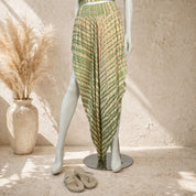 A mannequin wearing a green and beige striped skirt with a high slit, standing next to a pair of sandals and a vase of dried flowers.