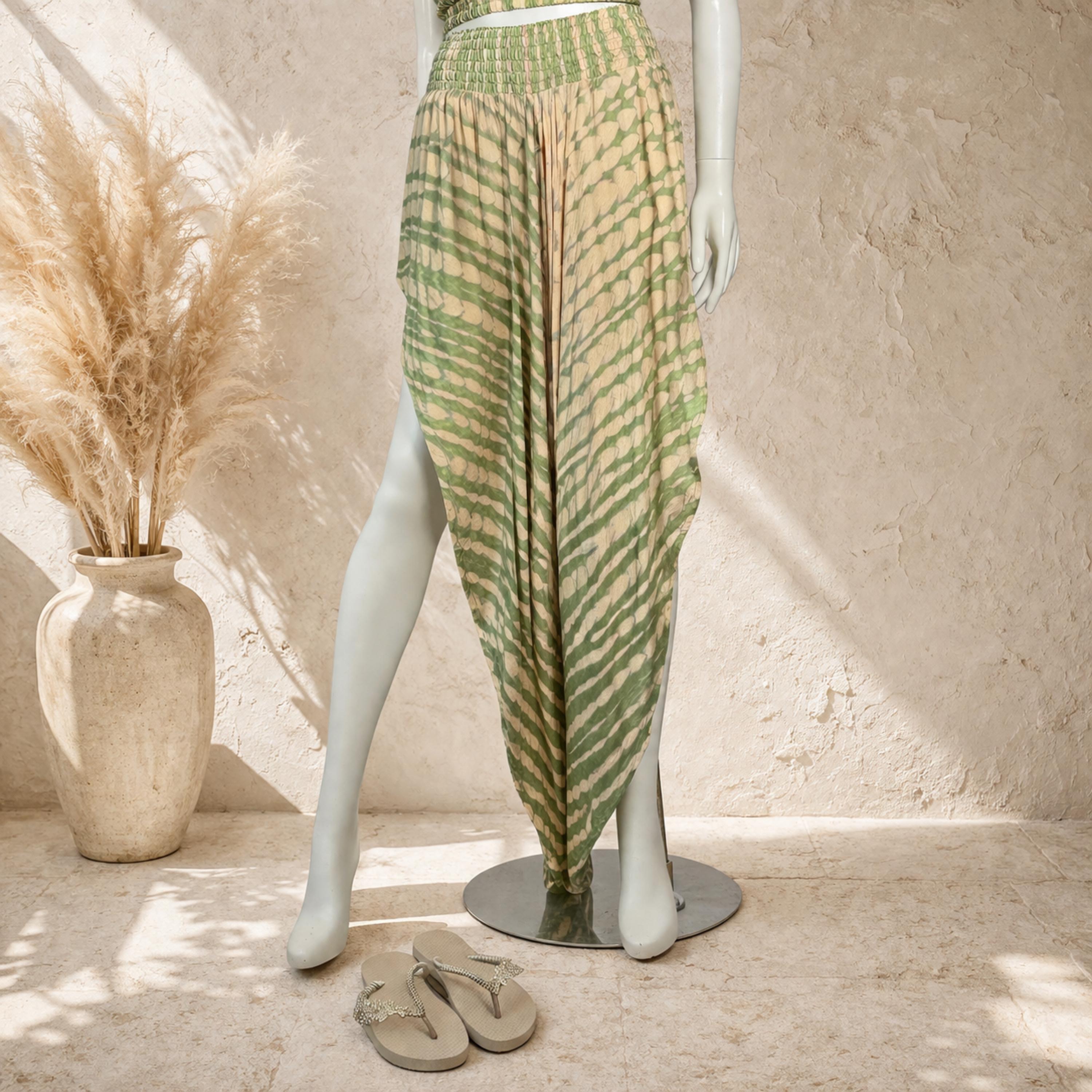 A mannequin wearing a green and beige striped skirt with a high slit, standing next to a pair of sandals and a vase of dried flowers.
