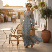 A woman in a blue floral dress stands in front of a white building, holding a woven purse and standing near a table with a cup and saucer.