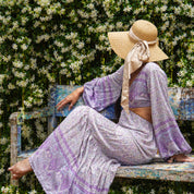 A woman in a long purple dress and wide-brimmed hat sits on a weathered wooden bench, surrounded by a lush green hedge with white flowers.
