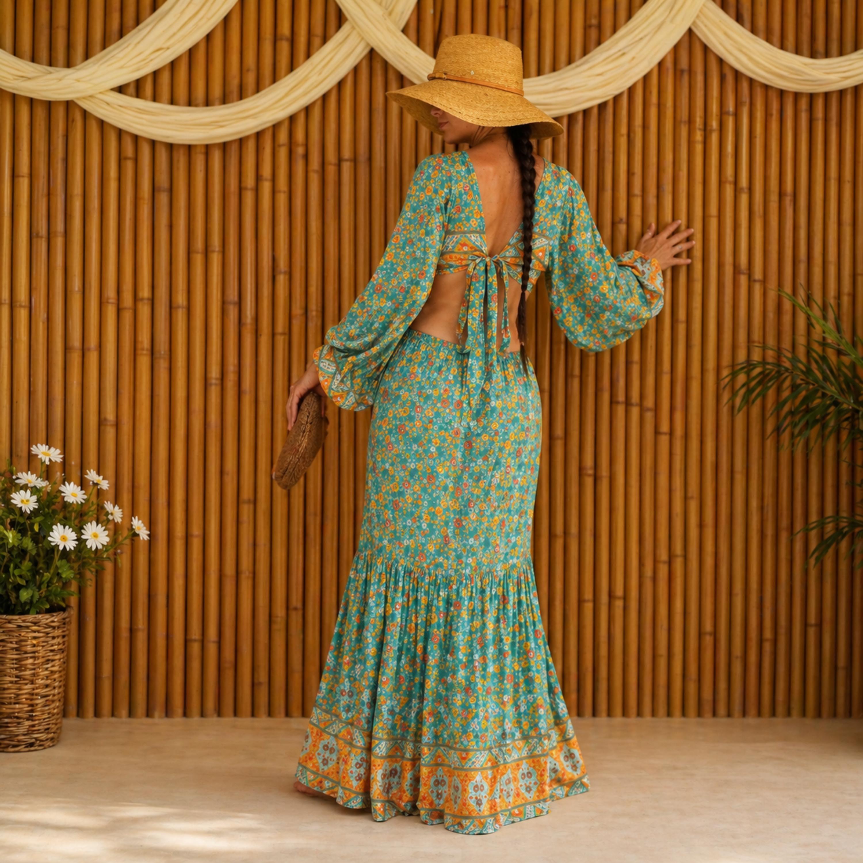 A woman wearing a colorful floral dress and a wide-brimmed hat stands in front of a bamboo wall, holding a small purse.