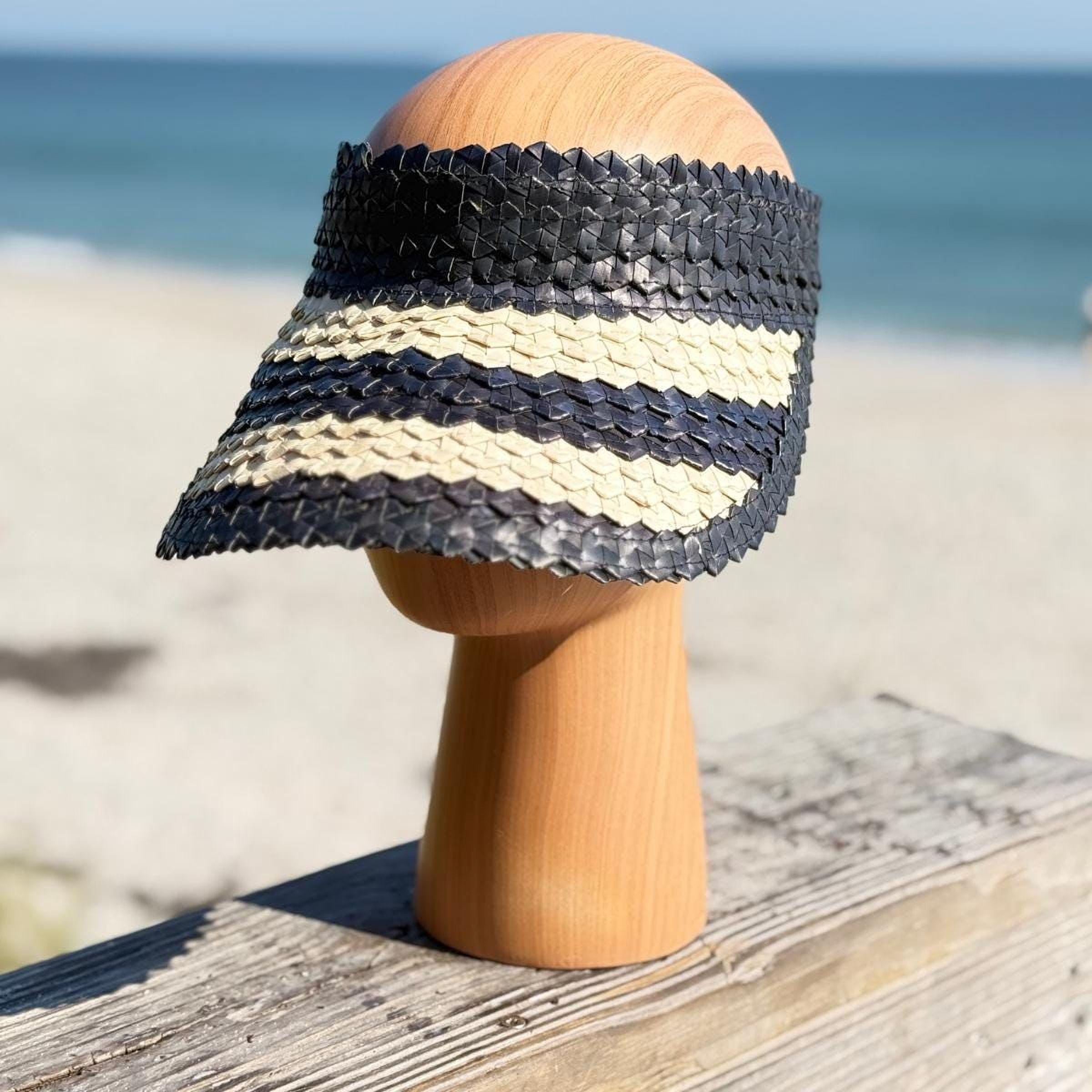 A wooden headless mannequin head wearing a black and white striped sun visor stands on a wooden surface, with the ocean visible in the background.