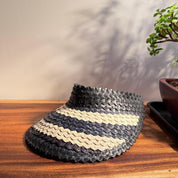 A woven black and white sun visor is placed on a wooden surface, with a potted plant and a wall in the background.