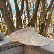 A white, textured visor is placed on a wooden surface, with a backdrop of trees and foliage.
