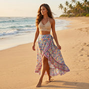 A woman in a beige crocheted top and a colorful floral skirt walks along a sandy beach, with palm trees and the ocean in the background.