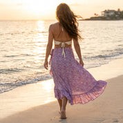 A woman in a purple floral skirt walks along a beach at sunset, with the ocean and a house visible in the background.