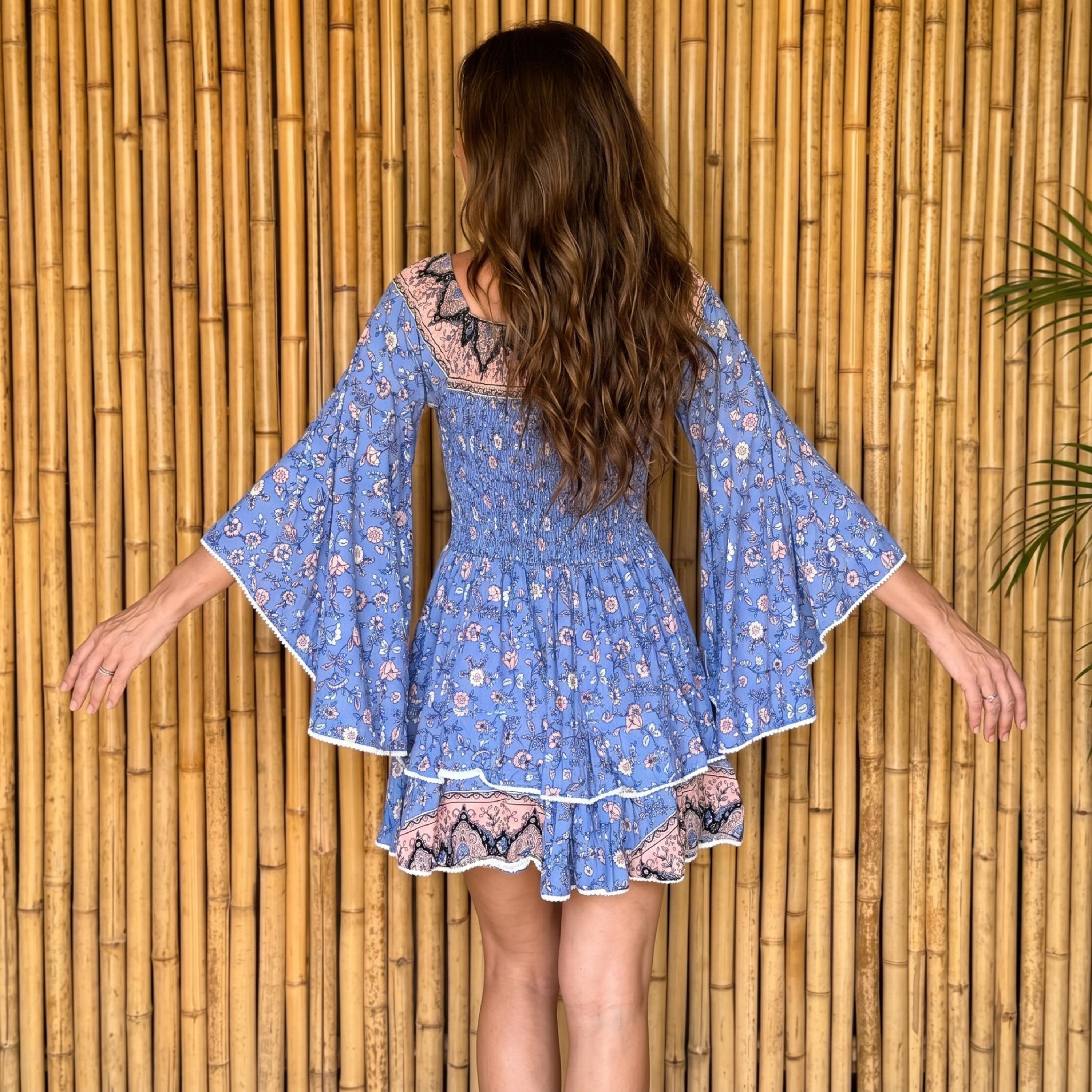 A woman in a blue floral dress stands in front of a bamboo wall, her back to the camera.