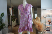 A mannequin wearing a purple floral wrap dress with a ruffled hem and a tie at the waist, displayed in a room with various decorative elements such as plants and a vase.