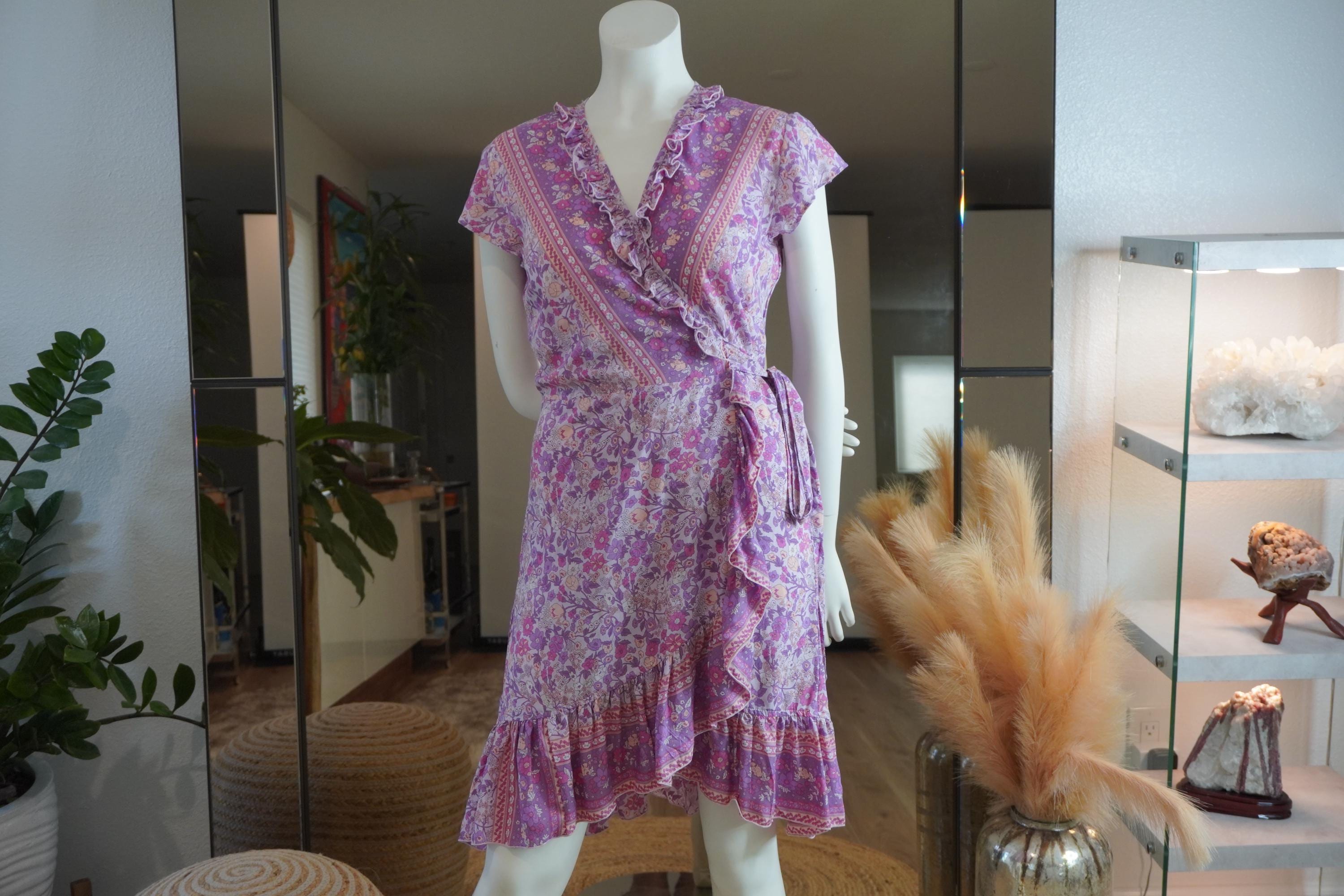 A mannequin wearing a purple floral wrap dress with a ruffled hem and a tie at the waist, displayed in a room with various decorative elements such as plants and a vase.