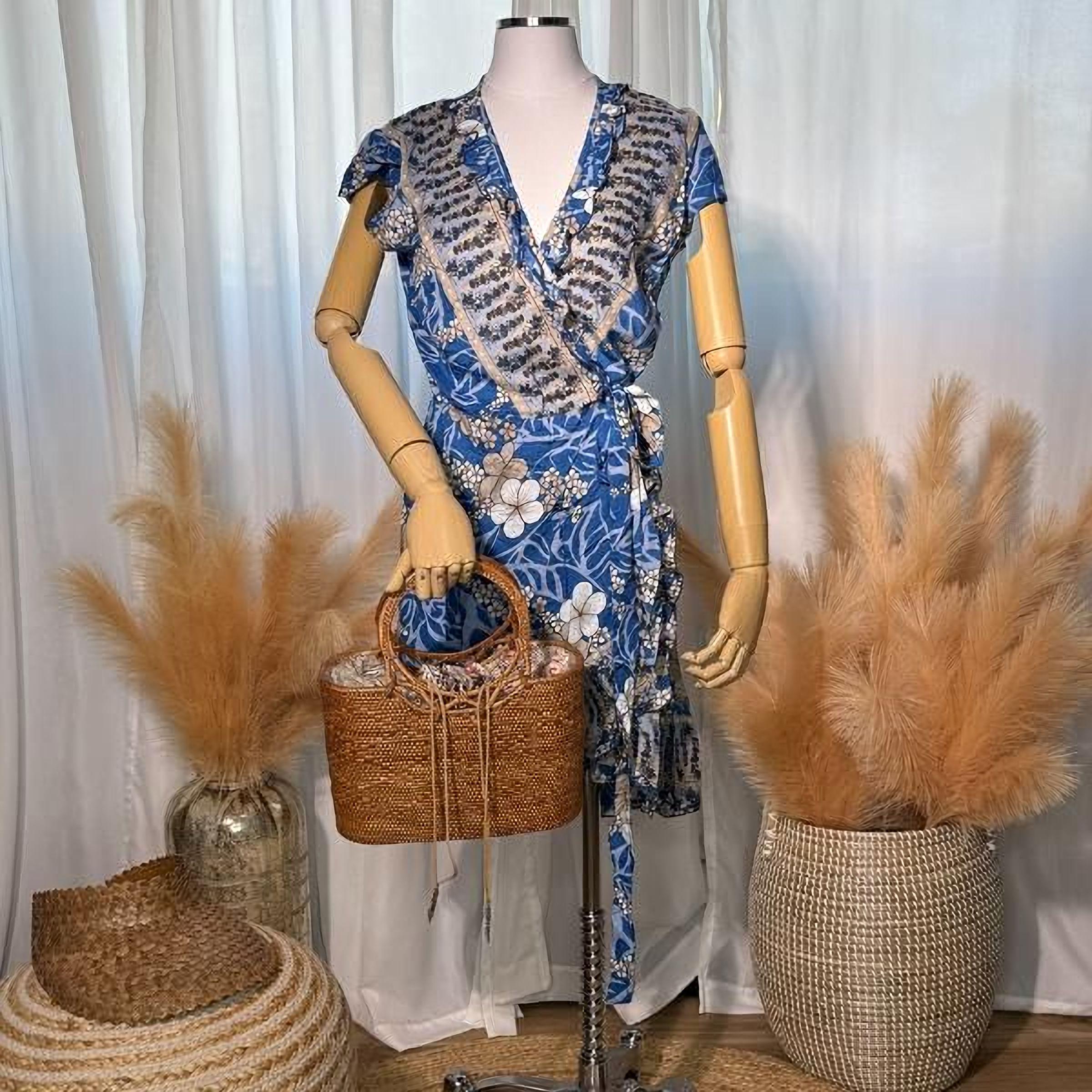 A mannequin wearing a blue and white floral wrap dress, holding a brown woven purse, with two large potted plants in the background.