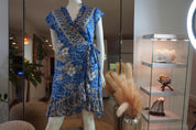 A mannequin wearing a blue and white floral wrap dress is displayed in a room with glass shelves and various decorative items.