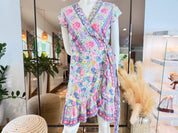 A colorful floral wrap dress displayed on a mannequin in a store, with a potted plant and other decorative items visible in the background.