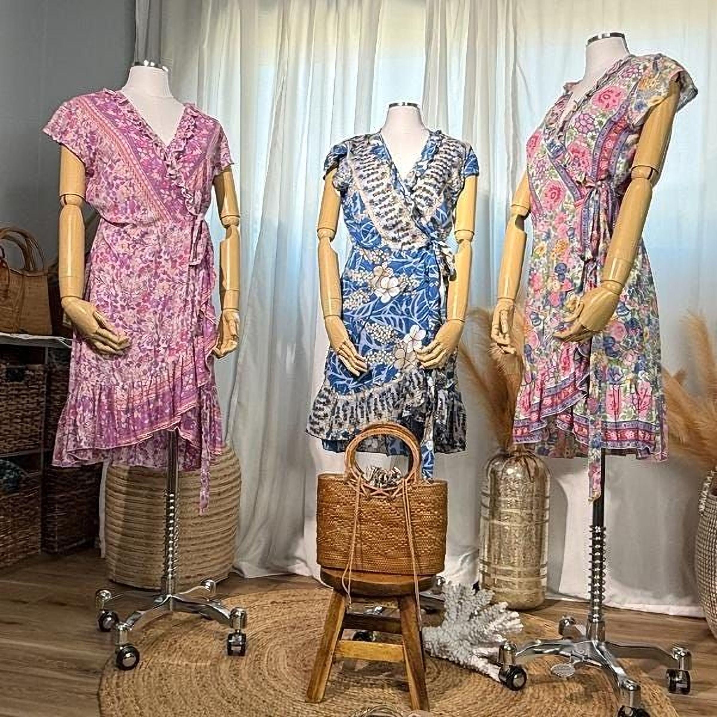 A display of three colorful dresses on mannequins, with a handbag and decorative elements in the background.