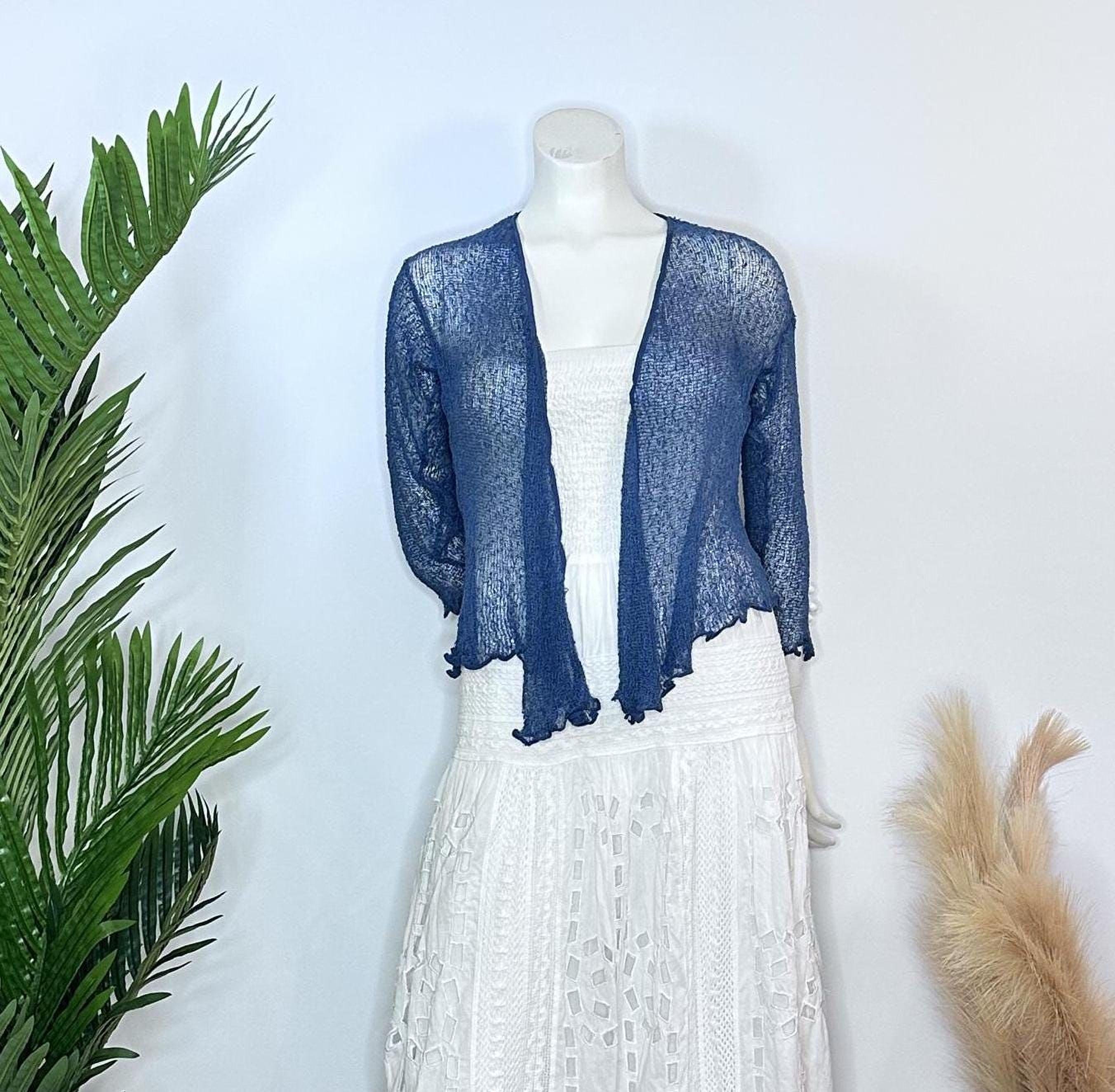A mannequin wearing a blue knitted cardigan over a white lace dress, with a potted plant to the left and a beige furry object to the right.
