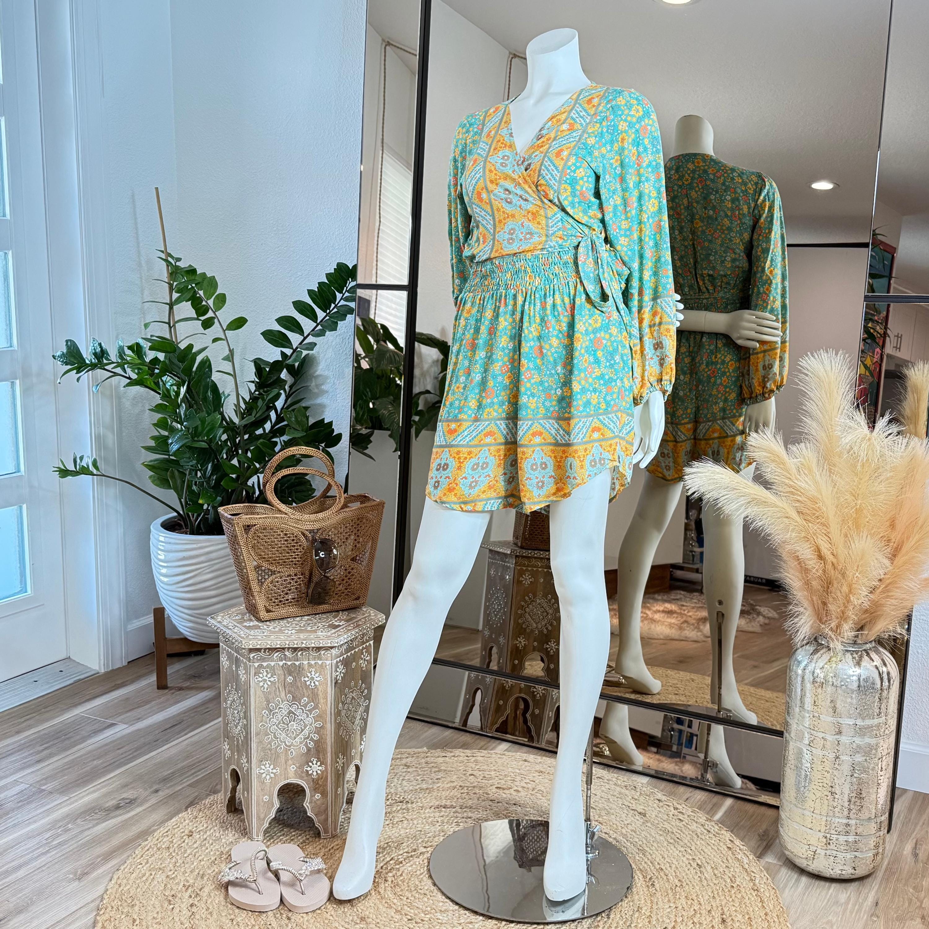 A mannequin wearing a colorful wrap-style dress with a floral pattern, placed on a wooden stool in a room with potted plants and a mirror.