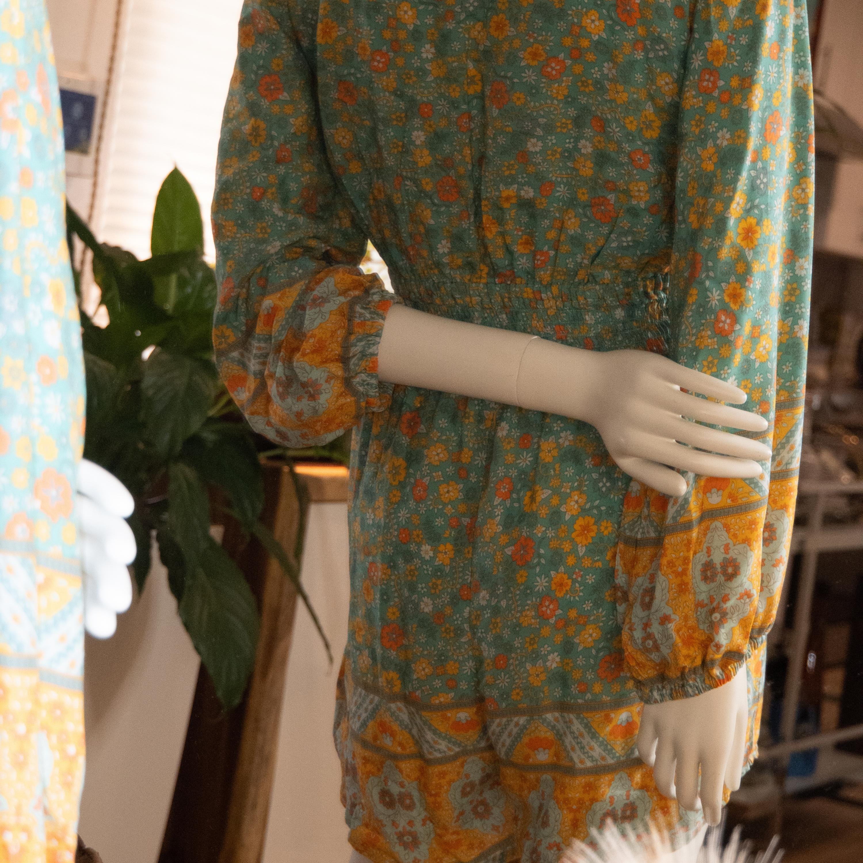 A mannequin wearing a colorful floral print dress stands in a room with a potted plant in the background.