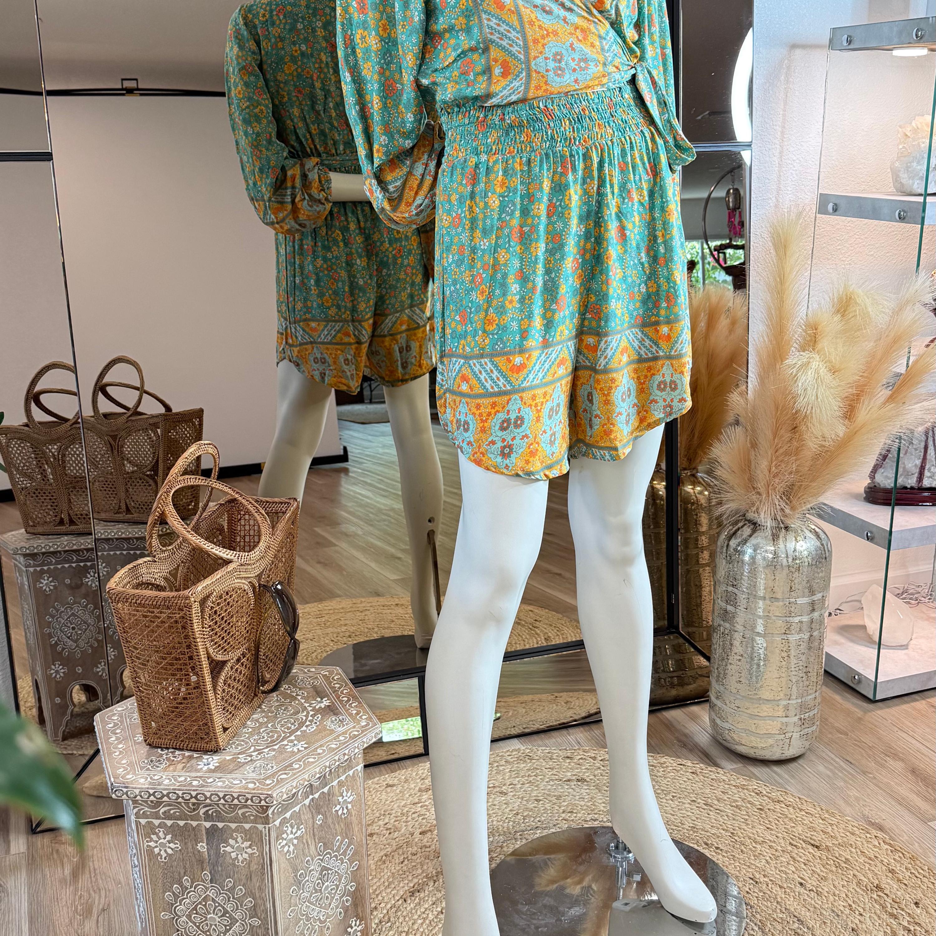 A mannequin wearing a colorful floral print dress stands in a clothing store, surrounded by various accessories such as a woven handbag and a decorative vase.