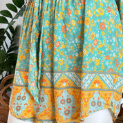 a colorful, patterned skirt with a floral design in shades of green, orange, and yellow. The skirt appears to be made of a soft, flowy fabric and is displayed against a backdrop of a plant.