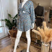 A mannequin wearing a blue floral print dress with a tie waist, placed in a room with potted plants and a mirror.