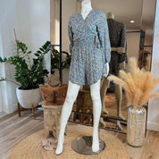 A mannequin wearing a blue floral print dress stands on a round rug in a clothing store, surrounded by potted plants and a mirror.