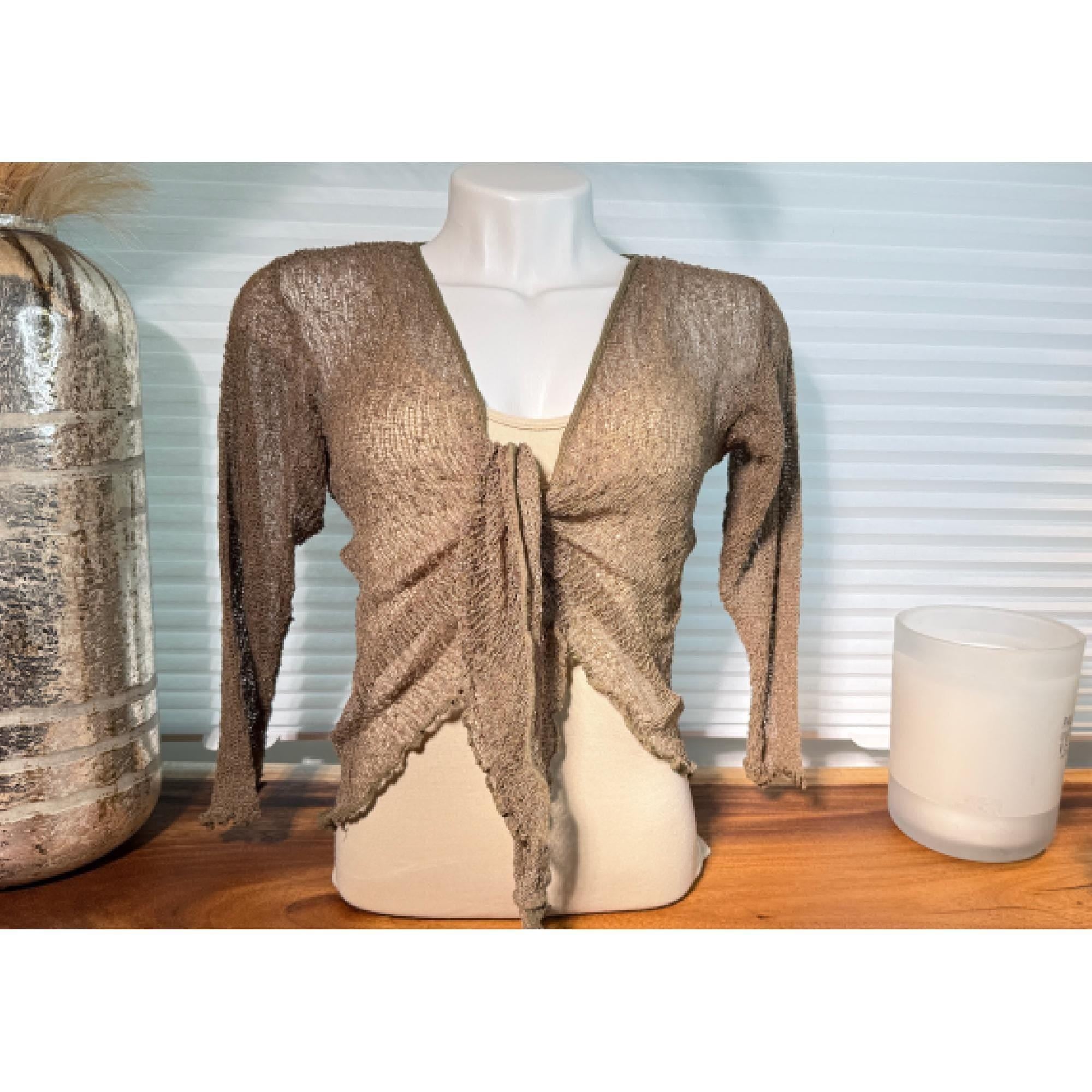 A mannequin wearing a beige knitted cardigan with long sleeves, displayed on a wooden surface next to a glass vase and a candle.