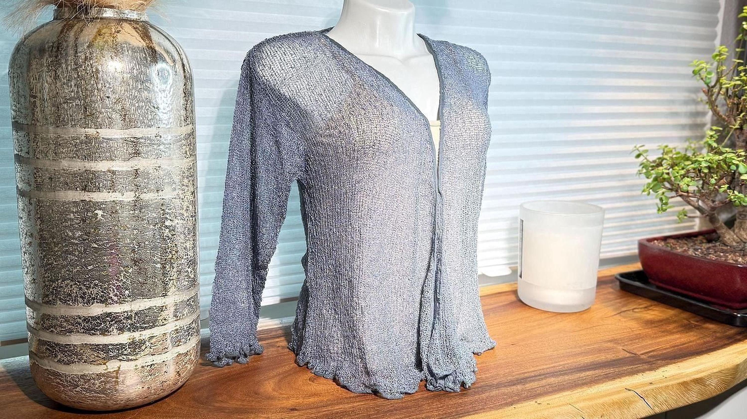 a mannequin wearing a blue knitted cardigan, placed on a wooden surface next to a large silver vase and a small potted plant.