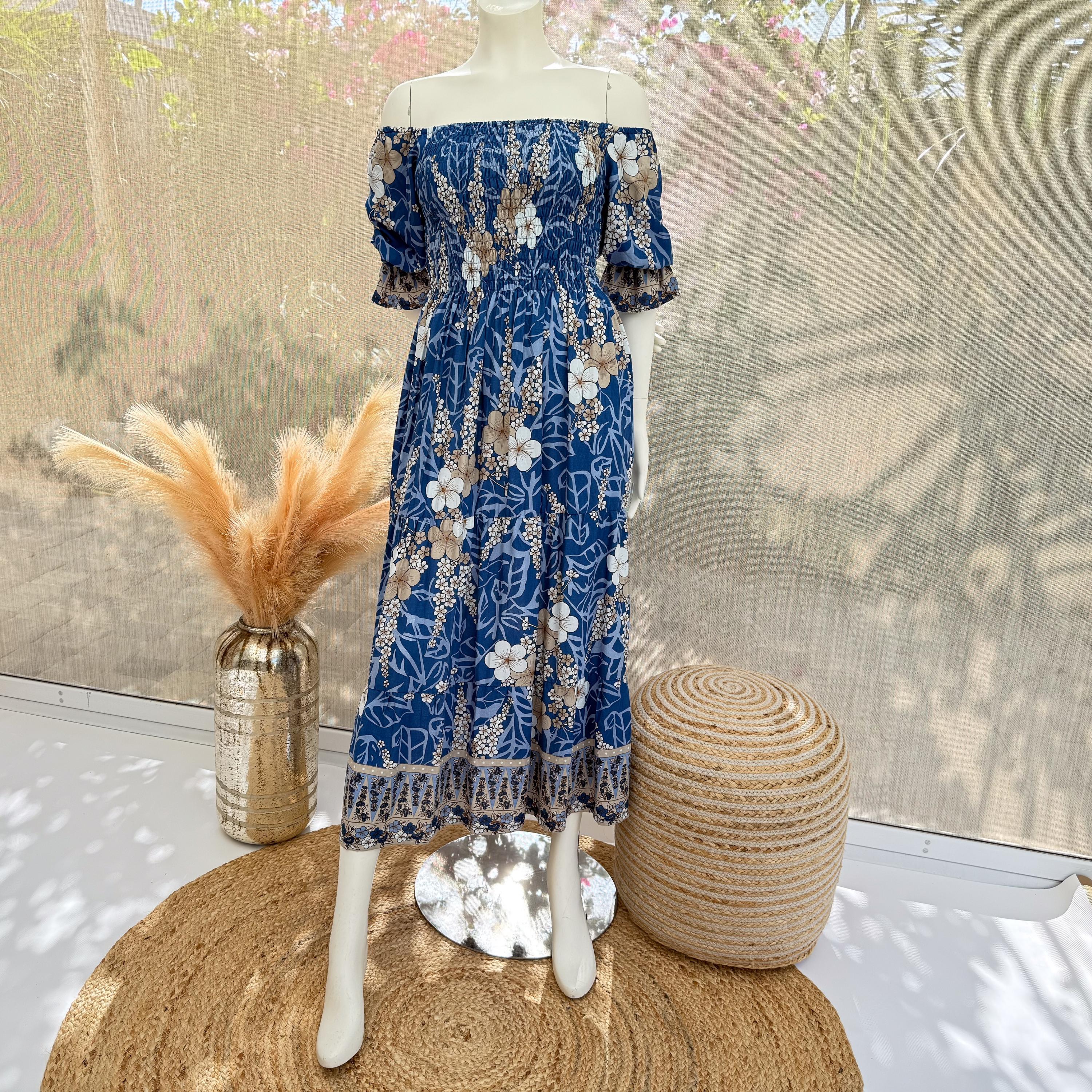 A mannequin wearing a blue and white floral dress stands on a round rug, with a woven basket and a vase of orange flowers nearby.