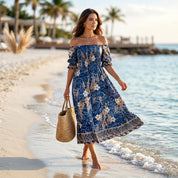 A woman in a blue floral dress walks along a sandy beach, carrying a woven straw bag.