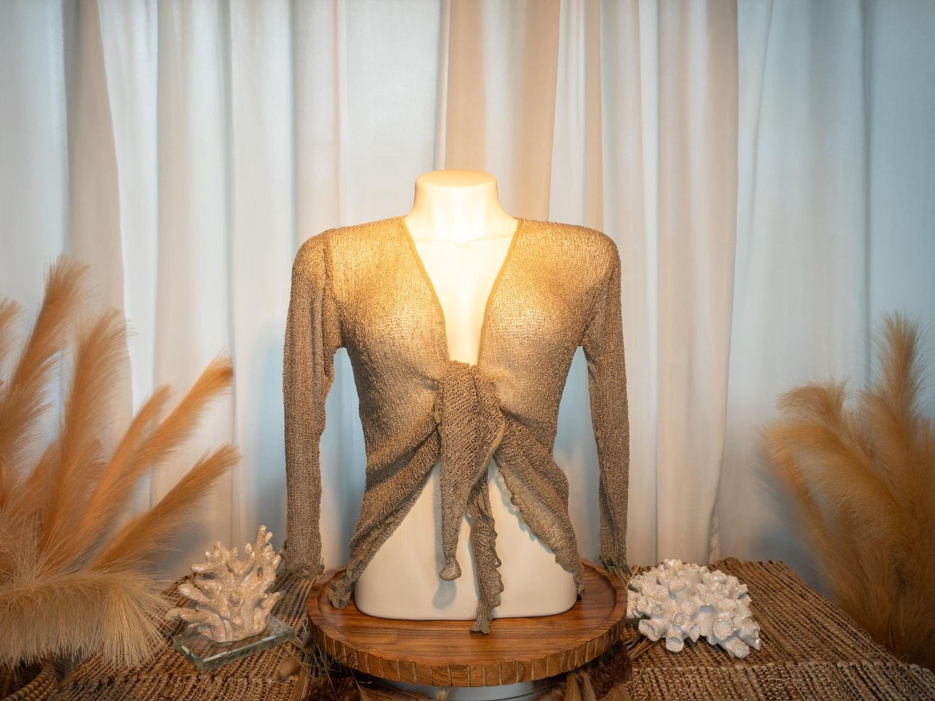 a mannequin wearing a beige knitted top with a tie detail, displayed on a wooden stand. The mannequin is positioned in front of a white curtain, and there are some decorative elements, such as a feather and a small white flower, placed around the mannequin.