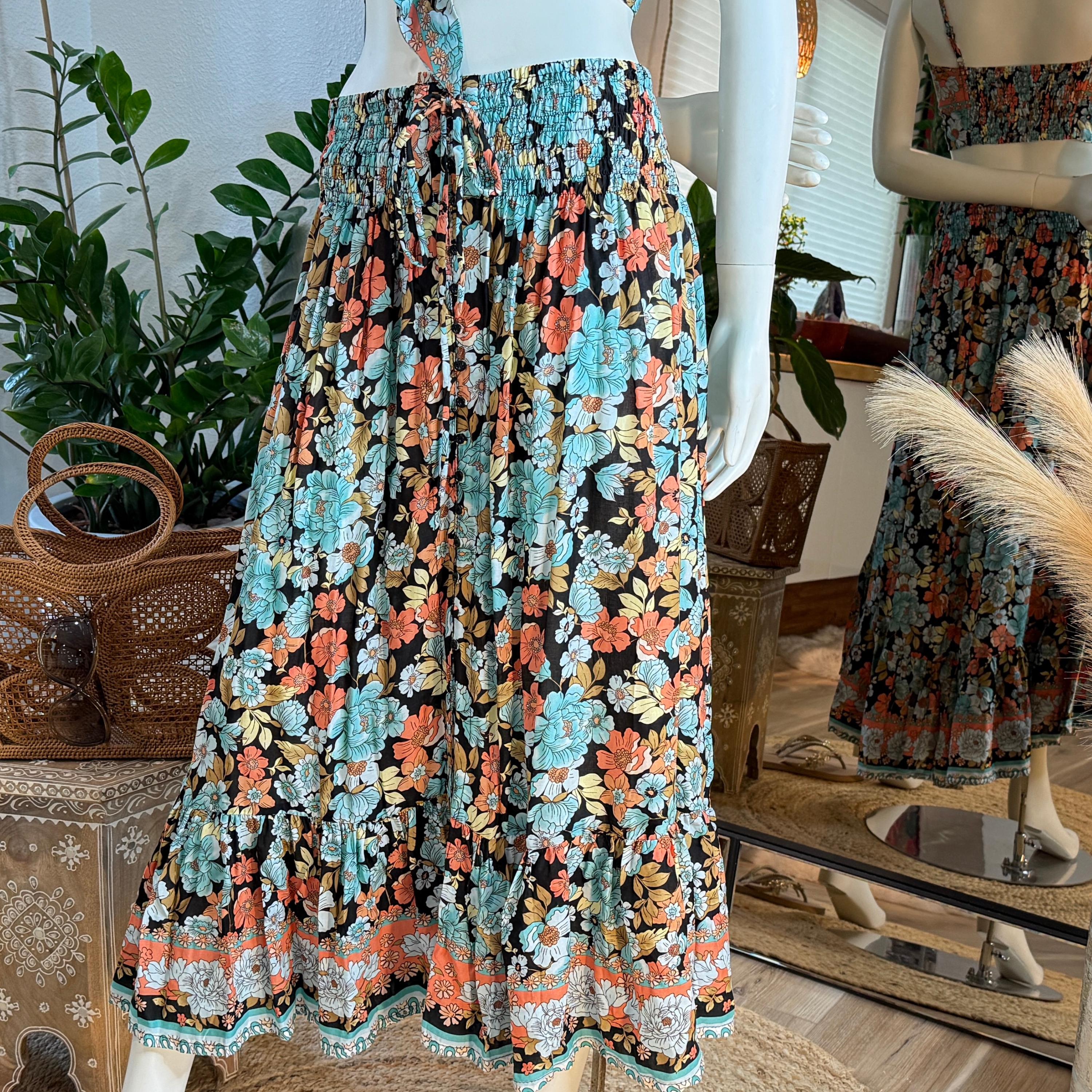 A mannequin wearing a colorful floral print skirt with a ruffled hem and a tie waist. The skirt has a mix of orange, blue, and green floral patterns. The skirt is displayed in a store setting, with potted plants and a mirror visible in the background.