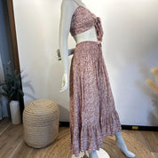 A mannequin wearing a pink floral print dress with a tie waist and a ruffled skirt, displayed in a retail setting with a woven stool and some plants in the background.