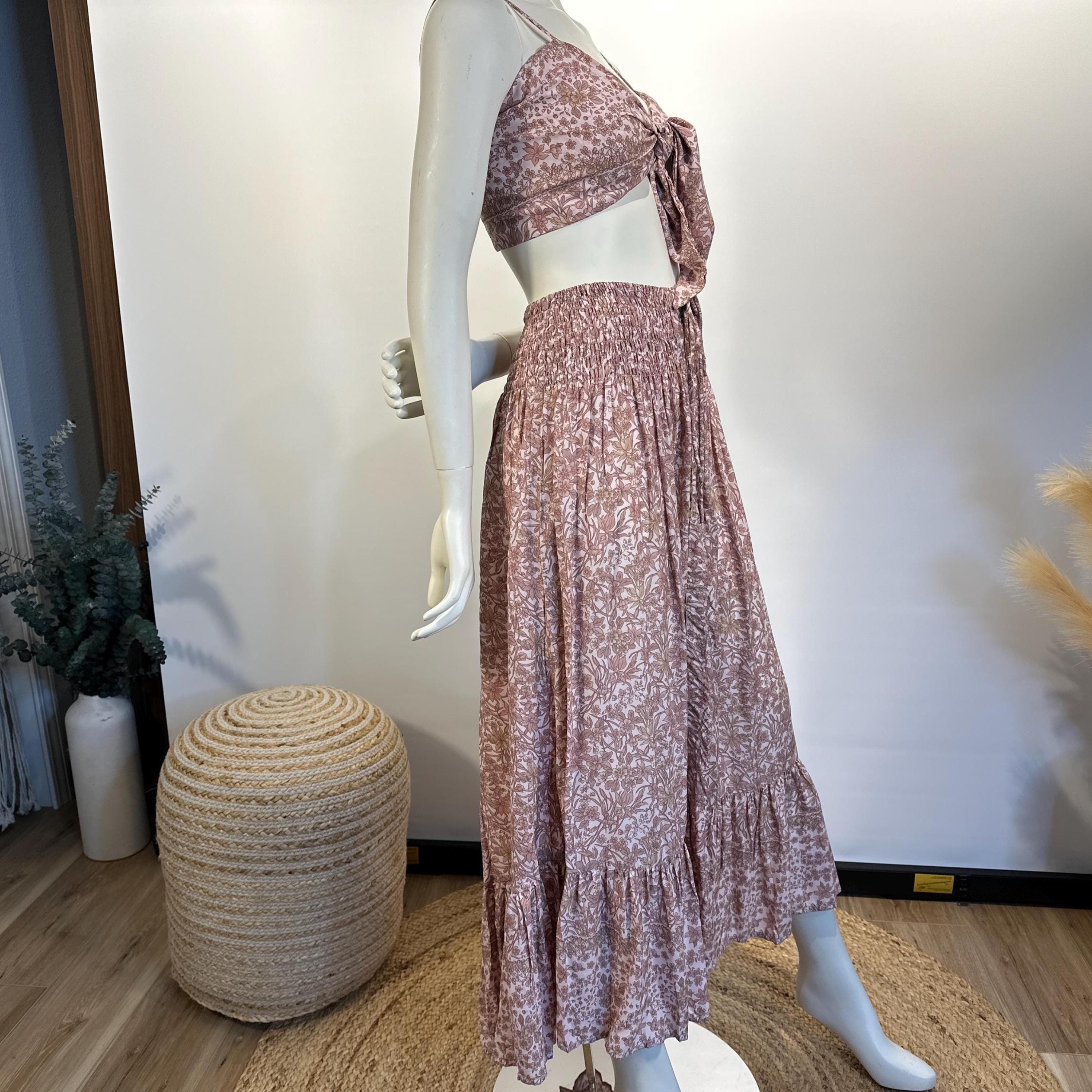 A mannequin wearing a pink floral print dress with a tie waist and a ruffled skirt, displayed in a retail setting with a woven stool and some plants in the background.