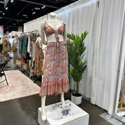 A mannequin wearing a colorful floral print dress and a matching top, displayed in a clothing store with various dresses and accessories visible in the background.