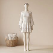 A mannequin wearing a white off-the-shoulder dress with bell sleeves stands in a room with a wicker basket and a pillow.