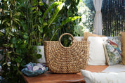 A woven straw handbag sits on a wooden table, surrounded by lush greenery and a cozy seating area with pillows and a bird-themed pillow.