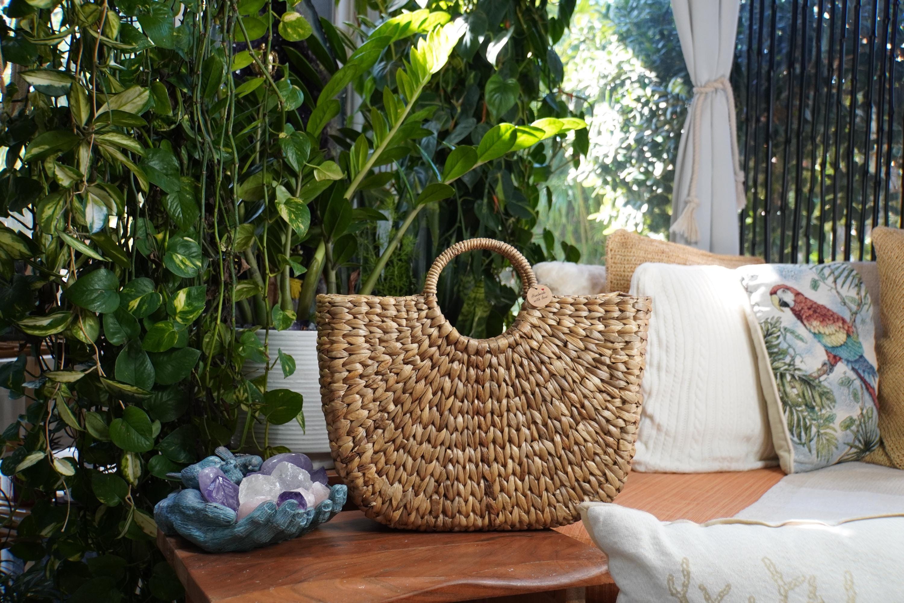 A woven straw handbag sits on a wooden table, surrounded by lush greenery and a cozy seating area with pillows and a bird-themed pillow.