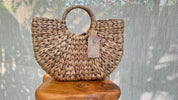 A woven handbag with a round handle and a tag attached, placed on a wooden stool against a backdrop of a white curtain.