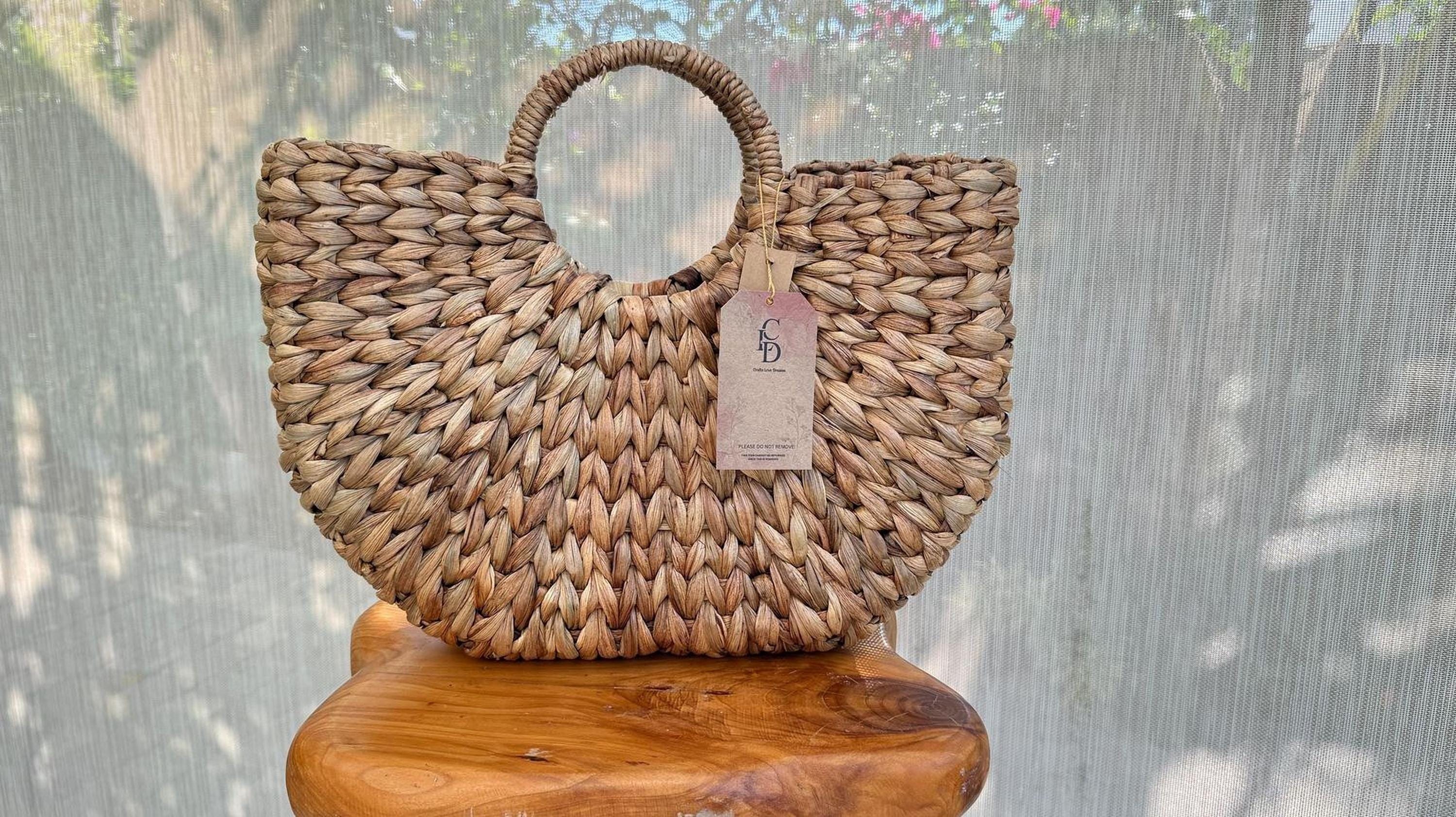 A woven handbag with a round handle and a tag attached, placed on a wooden stool against a backdrop of a white curtain.