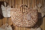 A woven straw bag with a handle, hanging on a bamboo wall.