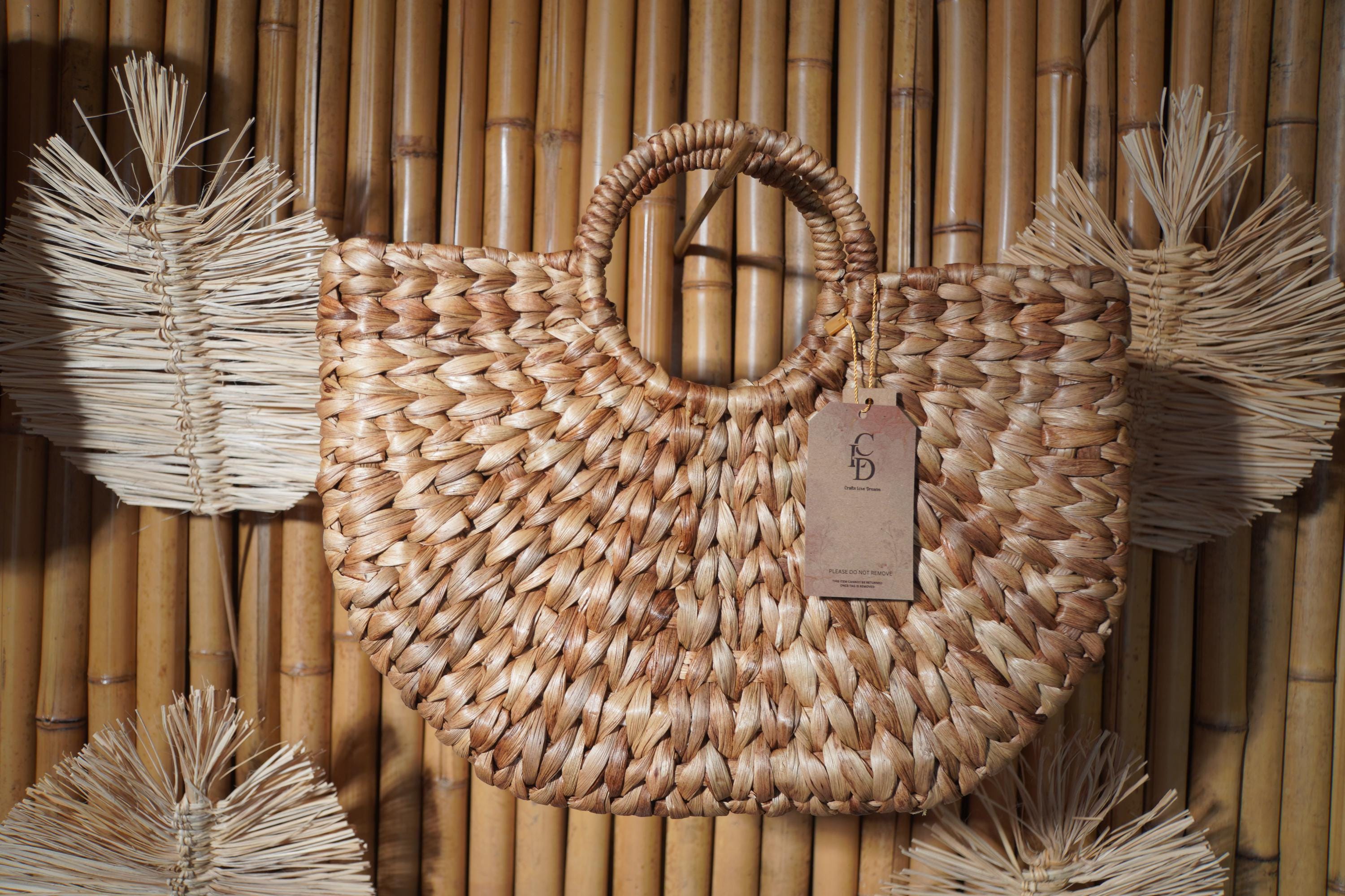 A woven straw bag with a handle, hanging on a bamboo wall.