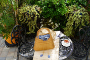 A woven straw bag rests on a wrought iron table, surrounded by potted plants and a white plate with a red drink, creating a cozy and inviting outdoor setting.