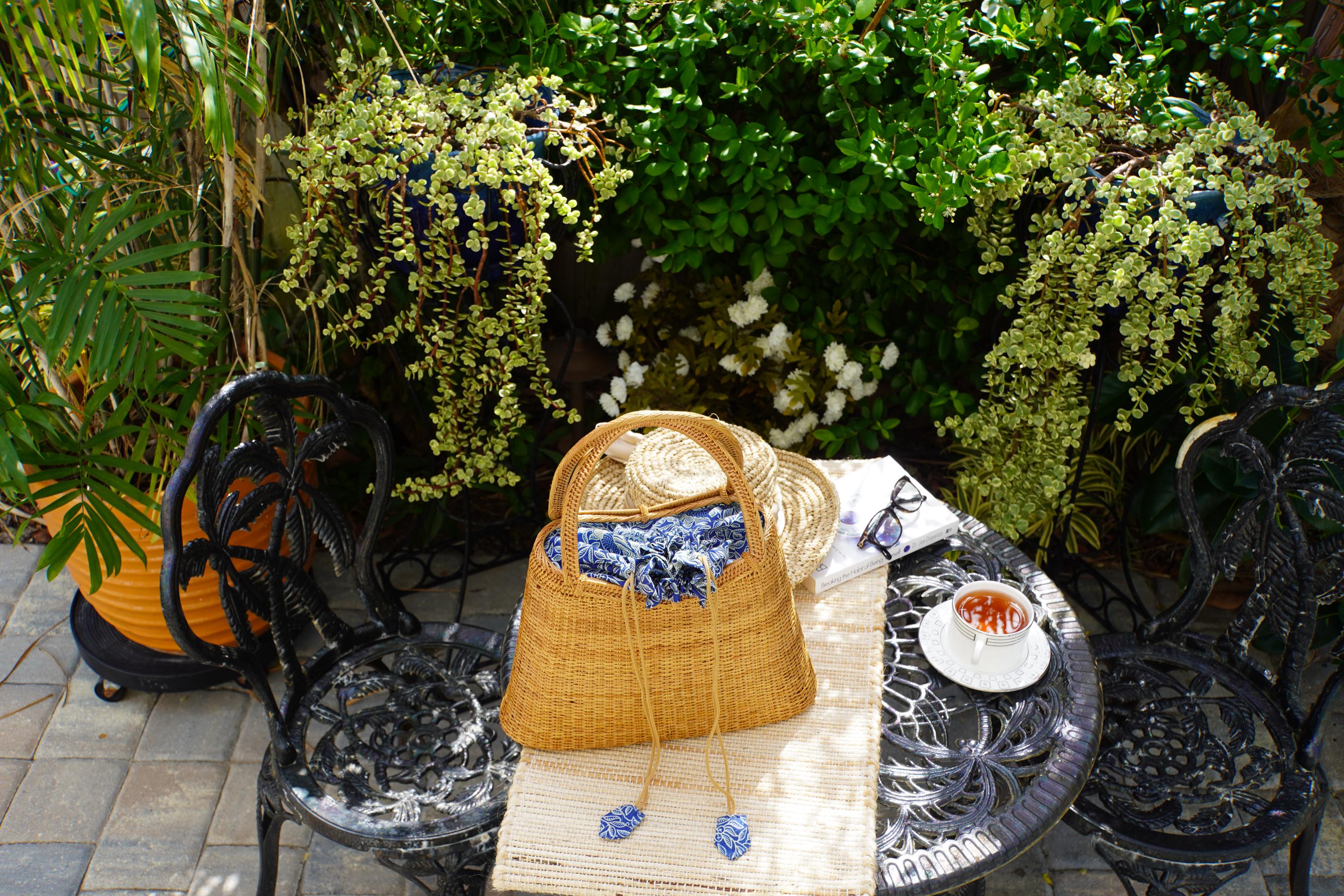 A woven straw bag rests on a wrought iron table, surrounded by potted plants and a white plate with a red drink, creating a cozy and inviting outdoor setting.