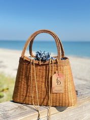 A woven brown handbag with a blue and white pattern sits on a wooden surface, with a tag attached to it, against a backdrop of a sandy beach and a clear blue sky.